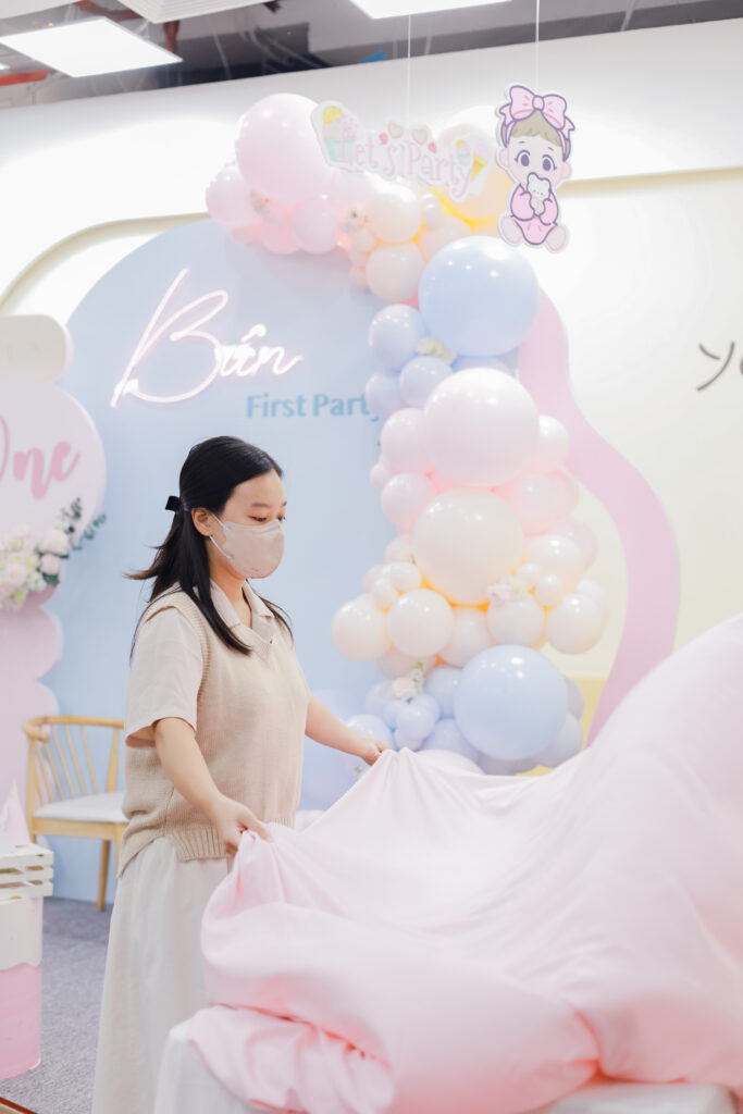 Nhân viên Mai Catering đang trải khăn bàn màu hồng pastel chuẩn bị cho tiệc sinh nhật bé, phía sau là background trang trí với bóng pastel và tên tiệc Bún First Party