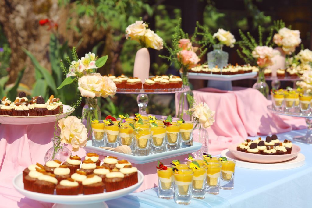Bánh cupcake và cheesecake xoài được bày trí trên khay sứ trắng giữa không gian tiệc sinh nhật ngoài trời với khăn trải bàn hồng pastel nhẹ nhàng