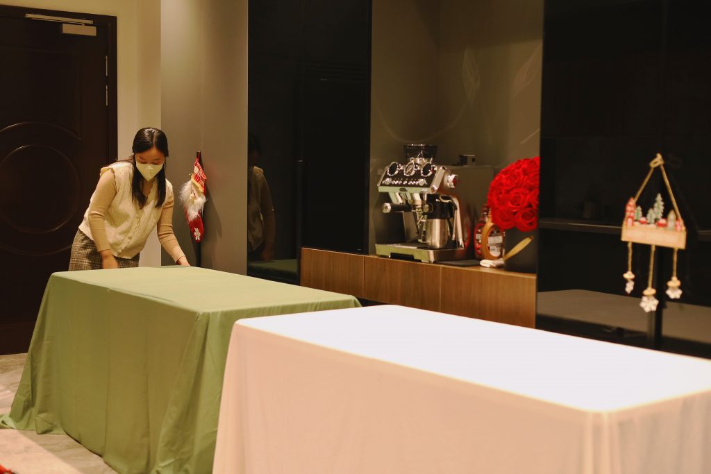 Nhân viên Mai Catering đang trải khăn lót trắng lên bàn tiệc để bảo vệ mặt bàn và tạo nền cho lớp khăn phủ chính, không gian chuẩn bị tại khu vực tiệc trong nhà